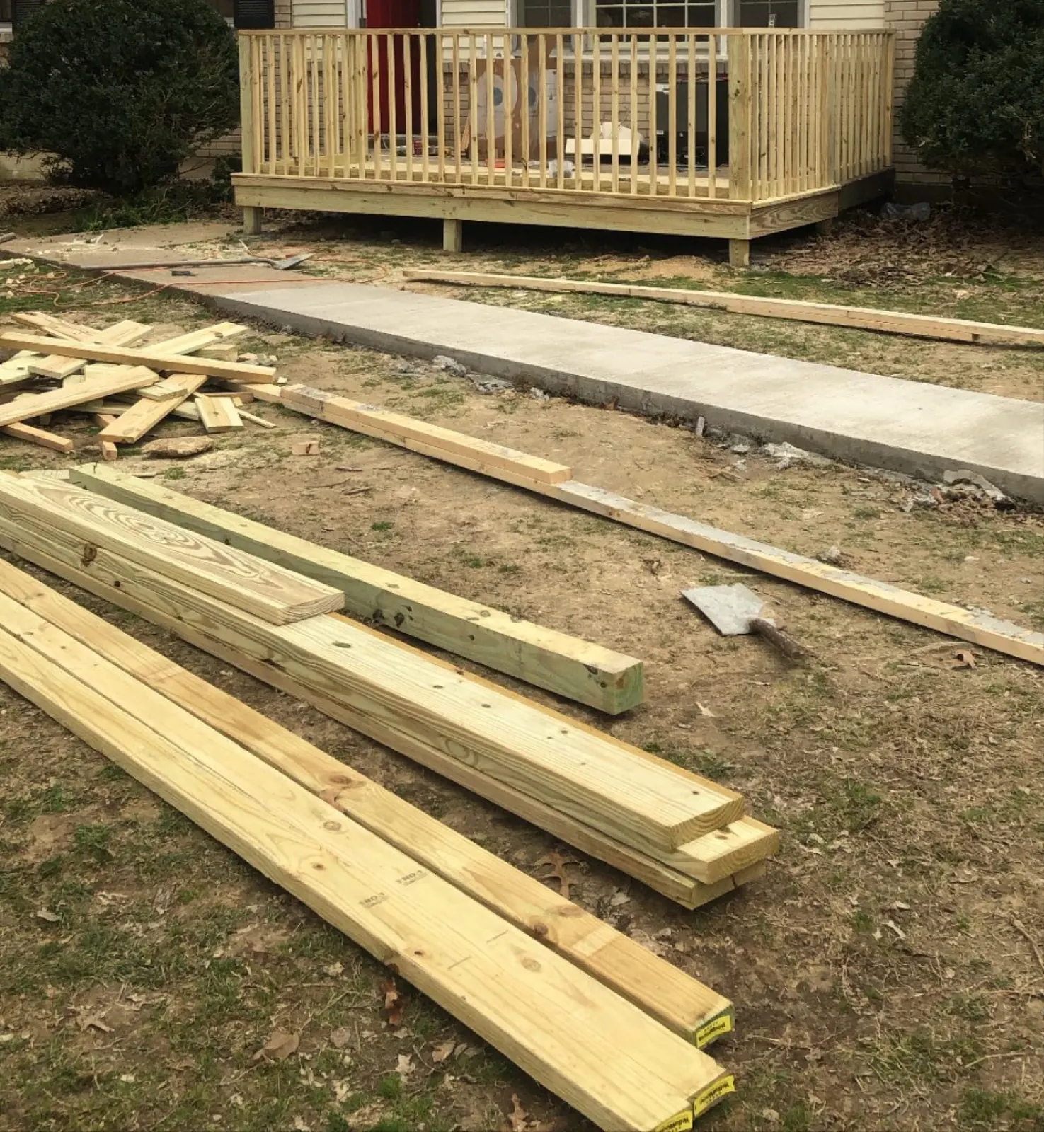 A new wooden deck with railings on the front of a house, with lumber stacked in the yard