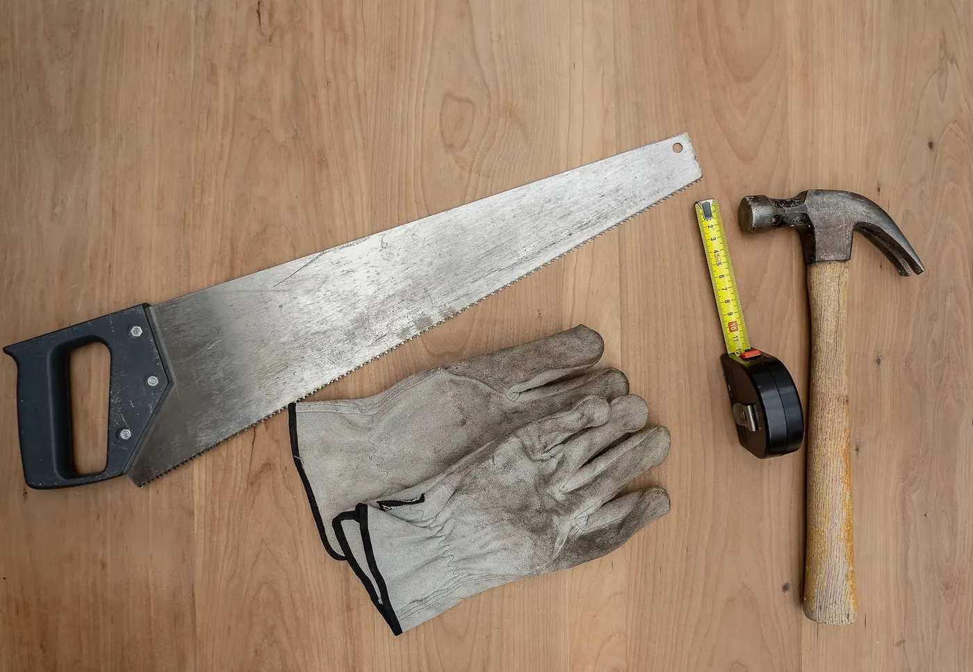 A handsaw, work gloves, tape measure and hammer laid out on a wooden surface